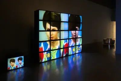 Five stacked rows of five monitors each, 1 monitor missing, display a woman, a single monitor on the floor repeats the image.