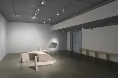 A concrete geometric bench sculpture sits on the floor, an object juts from a wall at back, a long bench lines the right wall.