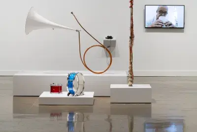 A collection of whimsical musical instruments are displayed on three white platforms. A video monitor displays a white male.