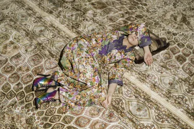 Woman in long floral-patterned dress reclines on and blends into the rich tan and red patterns of the carpet.