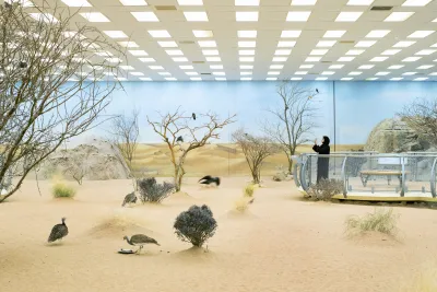 A person in hijab takes a photo in a gallery space with sand and blue painted walls.