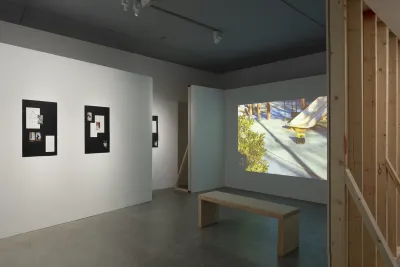 Projected color still of a playground and 4 paper works on black are shown on white walls with exposed wood framing.