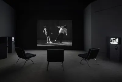 Photo of a black and white video projection and three chairs. Projection features a still of two children that appear to fall away from one another.