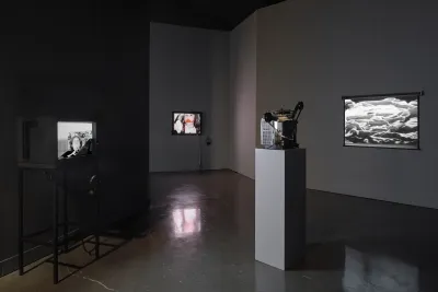 From left to right, a television set featuring a woman in black and white, a monitor featuring a young girl with lipstick smeared over her lips, and a projector and projection screen with black and white bedsheets.