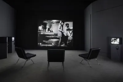 View from the back of the gallery with three large leather seats facing a black and white projection of two children playing in a messy room.