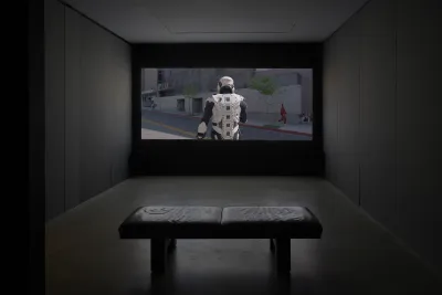 A dimly lit gallery space includes two video screens, one closer to the forefront on the left and one in the back right wall. A projector and a bench are also shown.