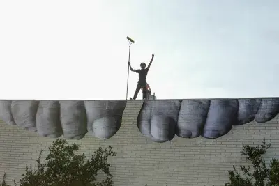 Artist JR stands on top of a building with a black and white at work on the side of a building.