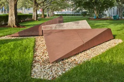 Corten steel sculpture surrounded by pebbles on a lush green grass.