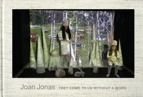 Book cover for Joan Jonas: They Come to Us without a Word featuring a large projector with an image of small houses and two females standing in front of the screen dressed in all white. There are tall cone sculptures around the two females and a sculpture of a rabbit.