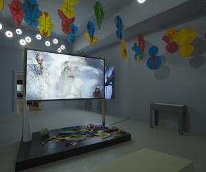 Installation view featuring a large screen in the center of the room playing a video and red, blue, and green shapes hanging from the ceiling.