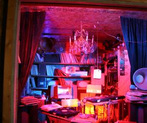 Installation view of a room inside a crate lit with red lights featuring cluttered albums and decorations.