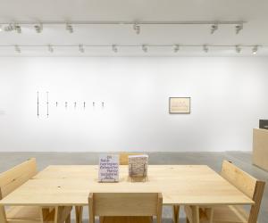 A white walled room with a tan wooden table and four tan wooden chairs in the middle of the room. To the right side of the table there is a wooden pedestal with a gray Plexiglas cube on top. There is a black framed drawing hung on the wall to the left of the pedestal. A row of eight test tubes are hung onto the wall to the left of the drawing. 