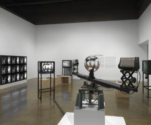 In the foreground, a sculptural apparatus comprises two cameras on the ends‬ ‭ of a single rotating arm, each pointed inwards at a central mirrored sphere. A feed from‬ ‭ the cameras are routed to a nearby monitor. In the background, other video monitors‬ ‭ are visible, including a grid of nine showing the same abstract black-and-white pattern,‬ ‭along with exhibition wall text.‬ ‭