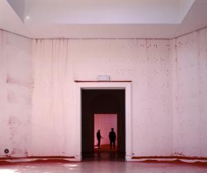 An open white-walled room with braille raised bumps across the walls. The silhouettes of two people can be seen through the dark doorway in the center of the image.