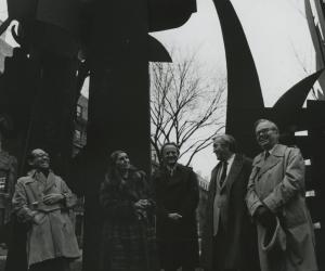 Black and white photograph of five people standing in a row outside in front of a tall black metal sculpture.