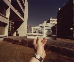 Photograph of a left hand holding up a rectangular photo of a black and white building. The photograph fits seamlessly with the street behind and surround buildings.