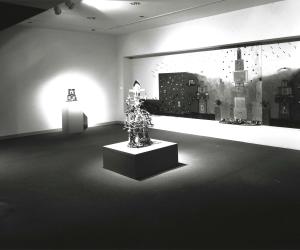 Wooden sculpture sits in middle of room, on walls are lit installation, colorful wall installation, and small sculpture.