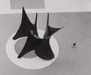 Black and white photograph of an aerial view os a large black abstract geometric sculpture. There is a person walking beside the sculpture.
