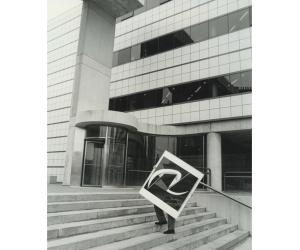 Black and white photograph of a person carrying a framed artwork up a set of cement stairs in front of a white building with dark windows that create a stripe pattern on the facade. The artwork is so large, only the person's legs are visible.