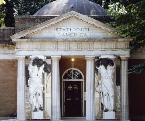 A classical stone building featuring tall columns supporting a grand entrance. There are two human figure statues on either side of the doorway. The text on the building facade reads, "STATI VNITI D'AMERICA"
