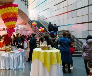 A crowd gathers in the List Center's lower atrium. Pink, yellow, and orange balloons in a cone-like structure dnagle from the ceiling over food on a table with a sequin table cloth. Other tables with yellow pink and orange table clothes are scattered throughout the space. 