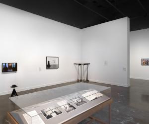 In the foreground, grayscale photographs and an open book occupy a wooden vitrine. The photographs show people in interior spaces, like a man sitting in a kitchen near a stovepipe and a child in a bath near his mother. Other artworks fill the background, including a droopy sculpture, a video monitor depicting a lecture, and a photograph of a procession.