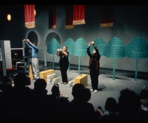 Three performers stand in front of the installation. With raised hands, they look up at a spotlight.