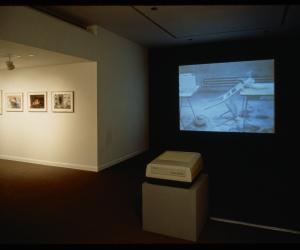 A projector shows a film in a dark gallery showing a chair resting against a table. Hanged works are lit on a wall nearby.