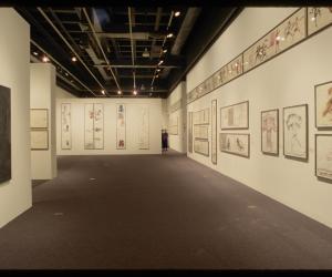 Thirty framed works on paper with colorful figurative sketches line the walls of a gallery. Two people stand in the distance.