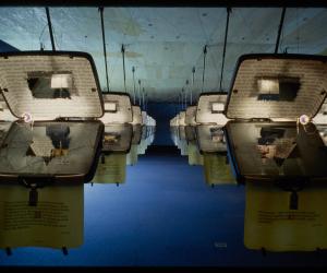Identical opened suitcases are hung from the ceiling in rows. Each has text projected on the inside. 