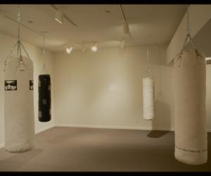 Three white and one black punching bags with text and images printed on them hang in four corners of gallery. 