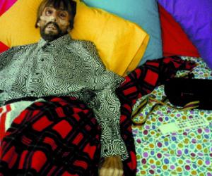  Photograph of bearded man with a sunken face on the edge of a bed covered in vibrant colored pillows and patterned sheets