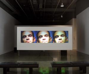 In a dimly lit space, a film screen displays 3 images of a man’s face, each image slightly different in coloration.
