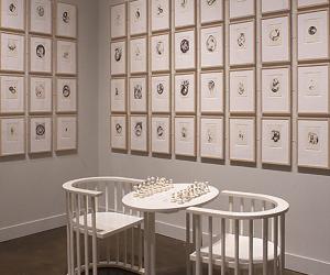 Installation of two white armchairs and a monochromatic chess table in front of a white wall, lined with framed prints 
