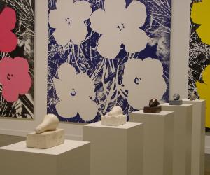 A series of lightbulb sculptures lie on pedestals, three large Warhol flower paintings hang on the wall in the background.