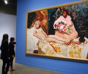 2 visitors view a large photo in a gold frame of a man portraying a nude woman lounging on a bed and a woman beside holding flowers. 