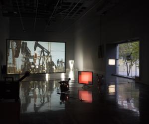 Projectors cast red rectangles on a small foreground screen, a large screen projection of oil drilling rigs in back.