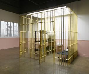 A yellow jail cell, with beds and a sink, stands in the middle of the space, while an image is projected on the wall to the left.