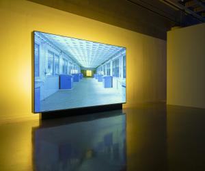 An image of a corridor with blue boxes and a patterned ceiling is projected on a standing screen backlit by yellow light.