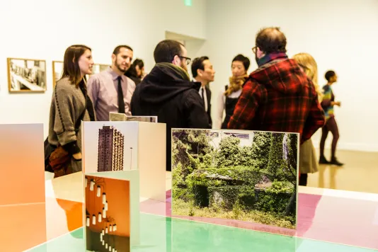 Members gather in a gallery. In the forefront is artwork that includes photographs of architecture and nature.