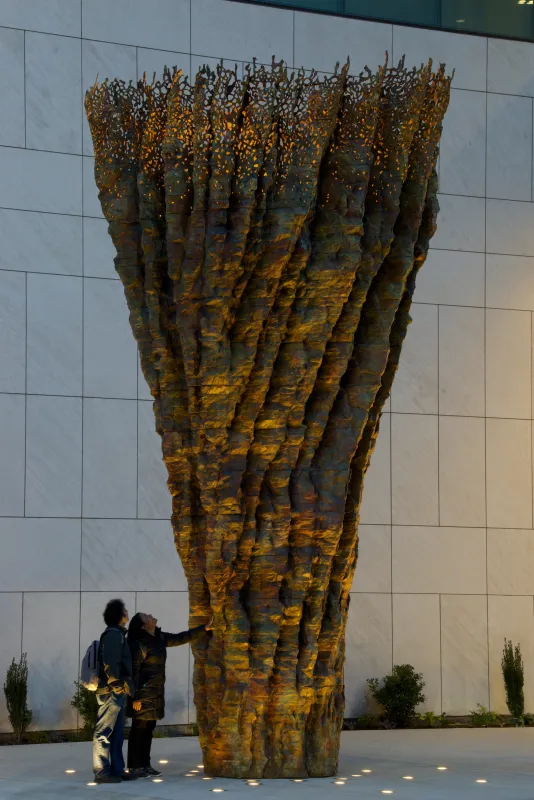 Bronze sculpture at night resembling a tree trunk with two people looking up at it on the sidewalk.