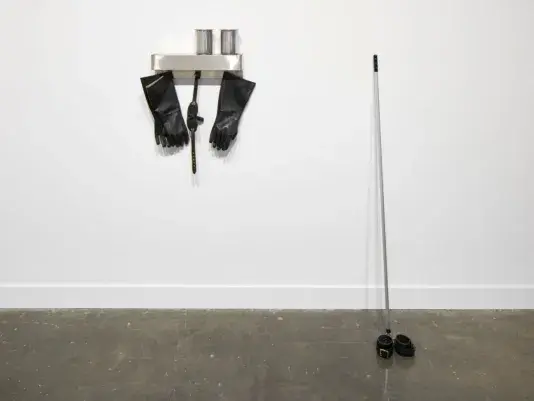 Metal pole leaning against the white gallery wall and to the left on a white shelf hangs a pair of black rubber gloves.