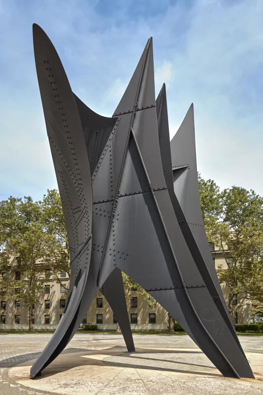 Installation view of large scale steel outdoor sculpture with a blue sky and buildings in the background.