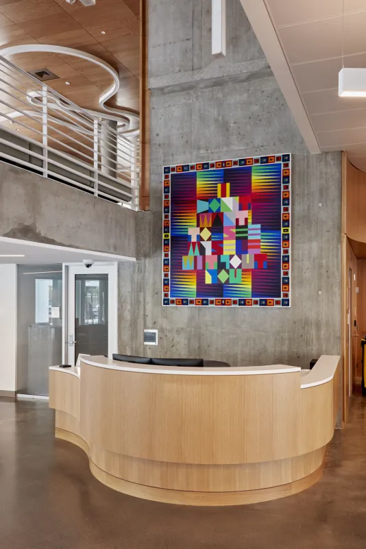 Installation view of a colorful painting hanging above a wooden desk in a lobby.