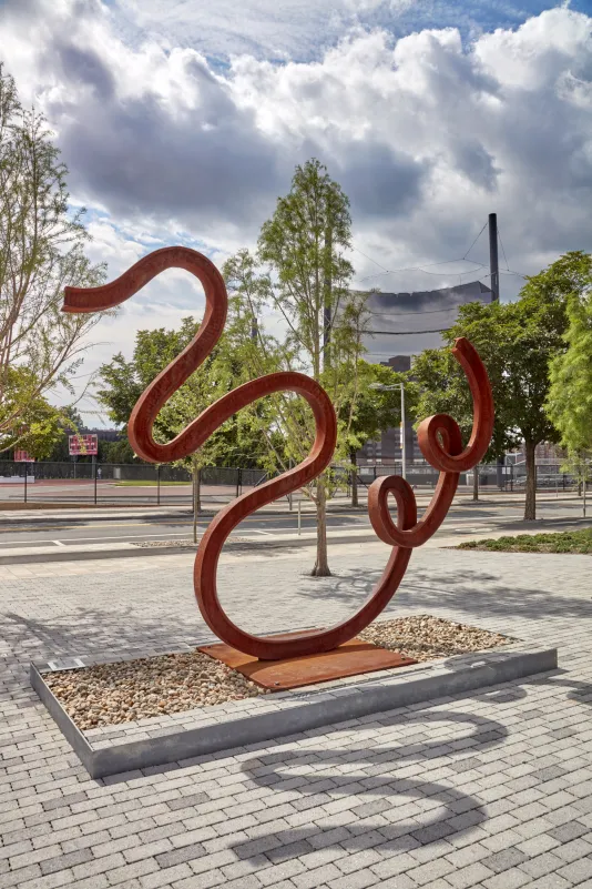 Matt Johnson's Untitled(Swan) sculpture of a warped bent metal train track is pictured in front of MIT campus buildings.