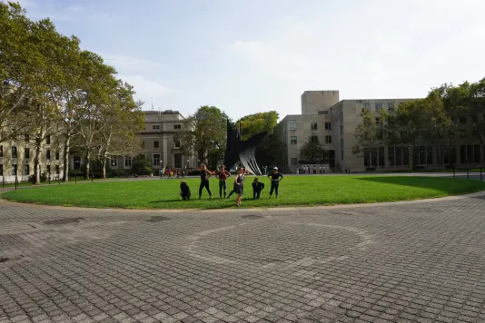 A group of people dance on a grass field in front of a steel sculpture.