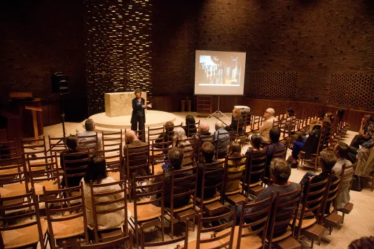 Woman stands at the front of a circular chapel with a projector screen and speaks to an audience who sits in wooden chairs.