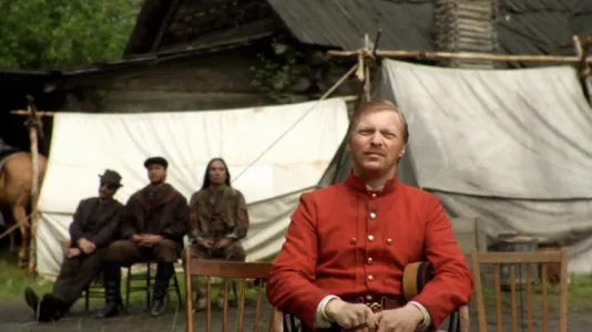 Man in a red shirt sits on a chair in the forefront with 3 people sitting behind him and white tents in the background.