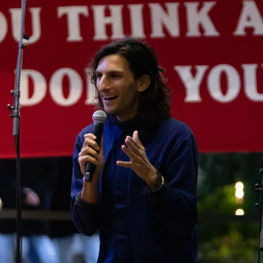 Artist Morgan Bassichis speaking into a microphone in front of a red sign.