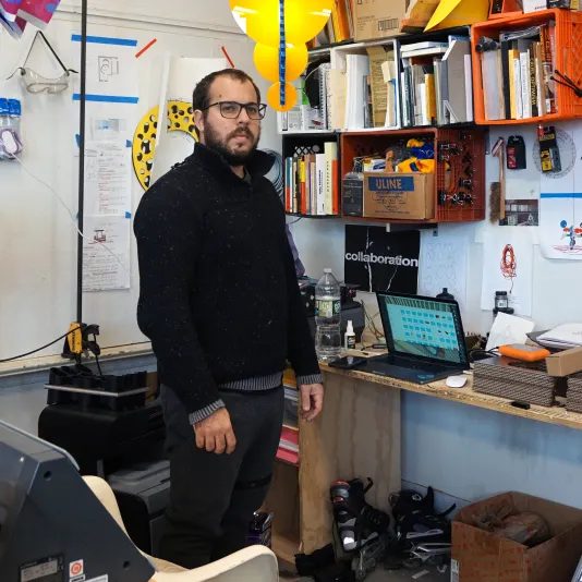 Rafael Domenech stands in his office surrounded by a books, a laptop, and paper hung on the wall.